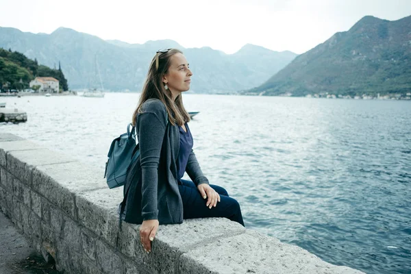 Mladá žena v šedou vestu a modrý batoh se nachází v blízkosti moře — Stock fotografie