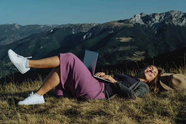 Jovem feliz viajante em um chapéu deitado na grama com laptop i — Fotografia de Stock