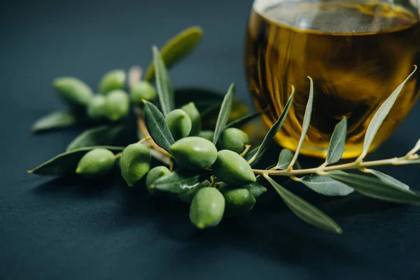 Aceitunas frescas, aceite en vidrio y rama verde sobre fondo negro — Foto de Stock