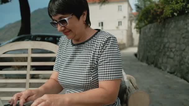 Mulher sênior imprime uma mensagem com laptop e sentado no café ao ar livre — Vídeo de Stock