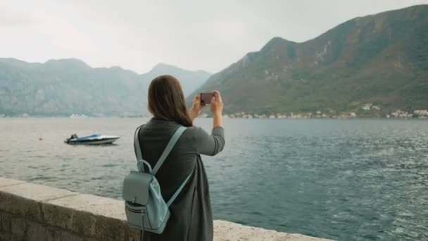Женщина в сером кардигане и голубом рюкзаке фотографирует горы по телефону у моря, Черногория — стоковое видео