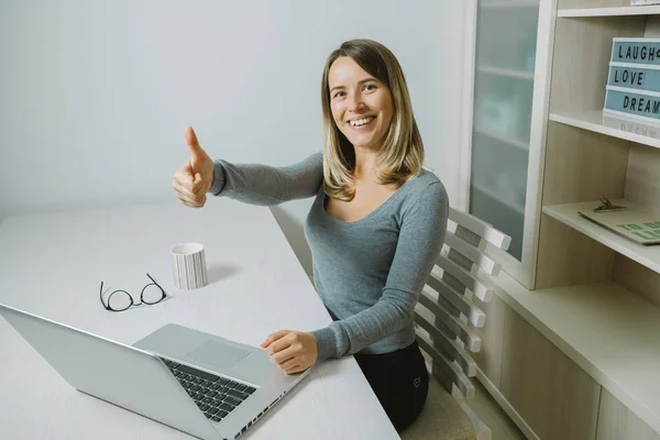 Smiling young woman thumbs up while working on laptop while sitt