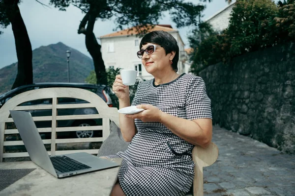 Leeftijd vrouw dranken cofee en zittend op openlucht café met laptop — Stockfoto