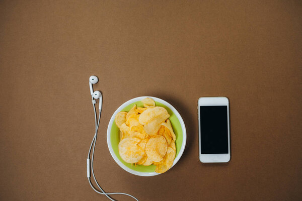 Top view of bowl with chips, phone and headphones on brown backgr
