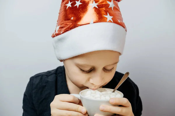 Симпатичный мальчик в красной шляпе и пьет какао в кружке. Рождественская концепция — стоковое фото