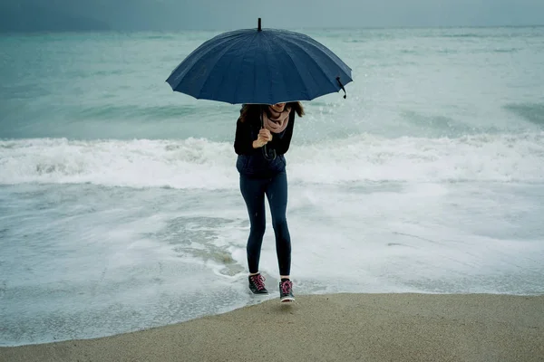 Веселая молодая женщина бежит от волны с зонтиком во рту — стоковое фото