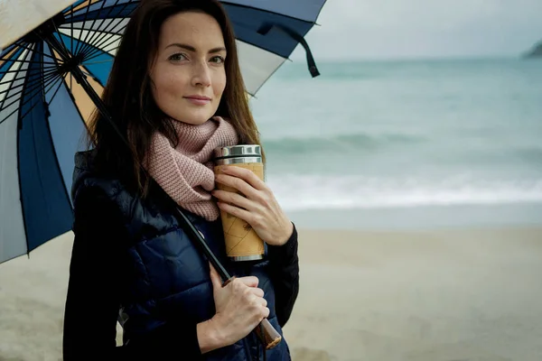 Jovem mulher de pé com um guarda-chuva e mantém uma garrafa térmica com h — Fotografia de Stock
