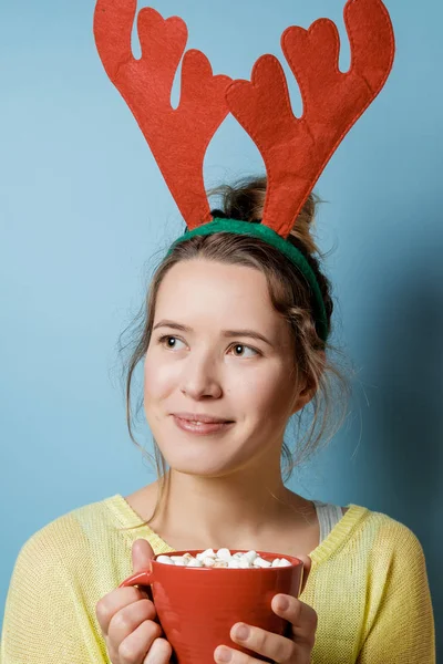 Schöne junge Frau in Weihnachtshörnern mit einem roten Becher auf einem blu — Stockfoto