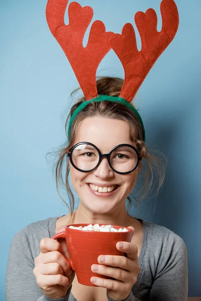 Schöne junge Frau in Weihnachtshörnern mit einem roten Becher auf einem blu — Stockfoto