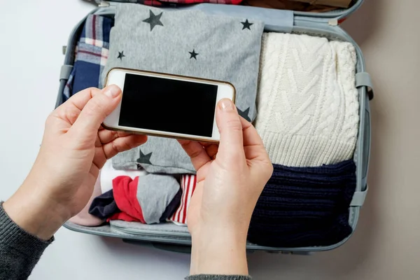 Manos de mujer con teléfono en el fondo de la maleta abierta lleno — Foto de Stock