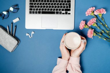 Düz yatıyordu kadın Ofis Masası. Kadın elleriyle kahve ve laptop, pembe karanfil, kırtasiye mavi masada. Üstten Görünüm