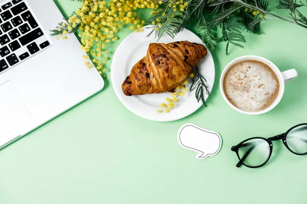 Woman work space with laptop, mimosa flower, croissant, coffee a