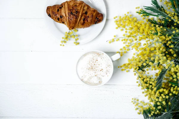 Schokoladencroissant und Cappuccino, dekoriert mit Mimosenblüten — Stockfoto