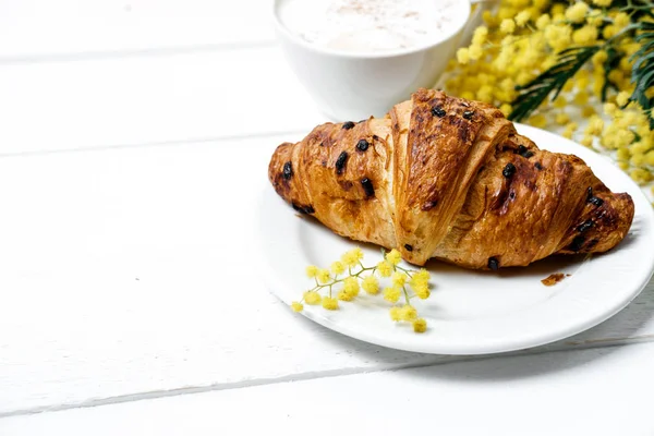 Choklad croissant och en cappuccino, dekorerad med mimosa-blomma — Stockfoto