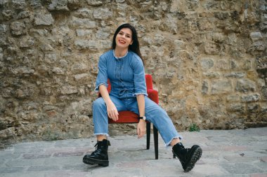 Kırmızı rujlu güzel, genç, gülümseyen esmer kadın arka planda bir koltukta oturuyor.