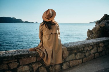 Bej ceketli, şapkalı, denize bakan ve gün batımını izleyen genç bir kadın..