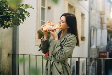 Yeşil ceketli güzel esmer kadın pizza yiyor ve şehirde dolaşıyor.