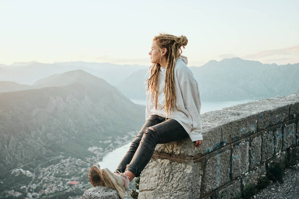 Smiling  traveler woman with dreadlocks  sitting on top of mountain and looking at sunset. Freedom concept