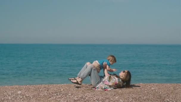 幸せな千年紀の女性は小さな男の子と遊んで 晴れた日に海のそばの海岸に座っています ママと息子は一緒に一日を過ごす — ストック動画