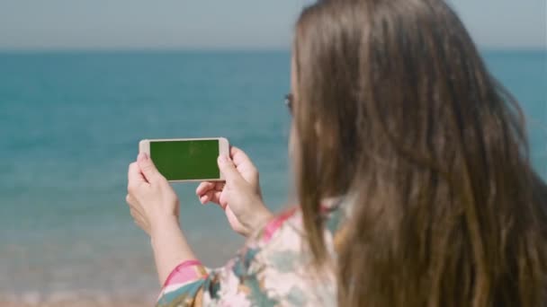 在阳光明媚的日子里 女性手紧握着装有绿色荧幕变色钥匙的智能电话机 站在海滩上 — 图库视频影像