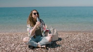 Metal bir şişeden su içen ve güneşli bir günde deniz kıyısında oturan genç bir kadın. Sıfır atık noplastik kavramı.