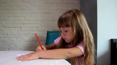 girl in a pink T-shirt sits near the window and is concentrated on drawing with a red pencil
