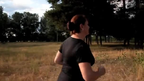 Mujer gorda en una camiseta negra corre a lo largo de un sendero forestal — Vídeos de Stock