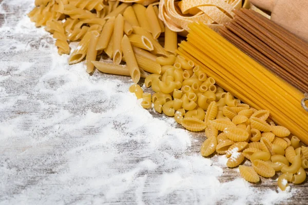 Different dry pasta on a wooden board with flour, top view — Stock Photo, Image