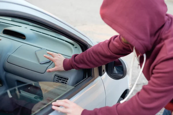 Hooded thief stealing a mobile phone from a parked car Royalty Free Stock Photos