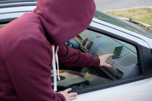 Ladrón encapuchado robando un teléfono móvil de un coche aparcado —  Fotos de Stock