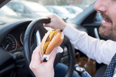 Adam arabasını sürerken hamburger yiyor.