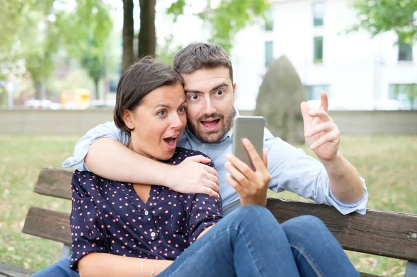 Casal feliz olhando para a tela do smartphone sentado em um banco — Fotografia de Stock