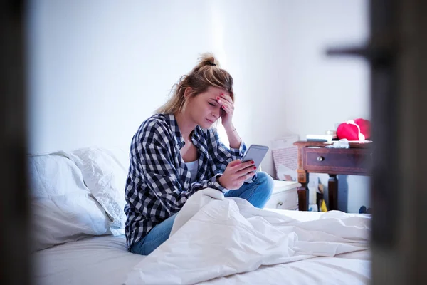 Молодая грустная эмоциональная симпатичная девушка, сидящая на кровати со смартфоном — стоковое фото