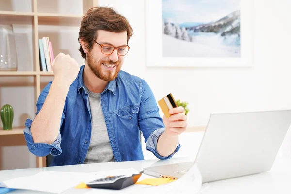 Sorrindo homem sentado em casa escritório e paga com cartão de crédito — Fotografia de Stock