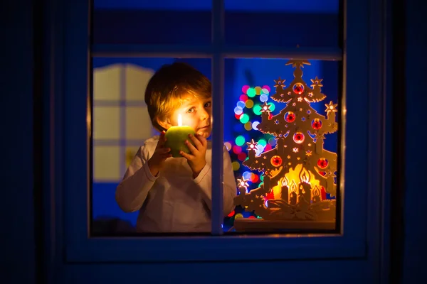 Little kid boy standing by window at Christmas time and holding — Stock Photo, Image