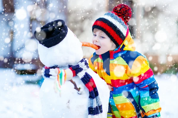 Funny kid boy in colorful clothes making a snowman, outdoors — Stock Photo, Image