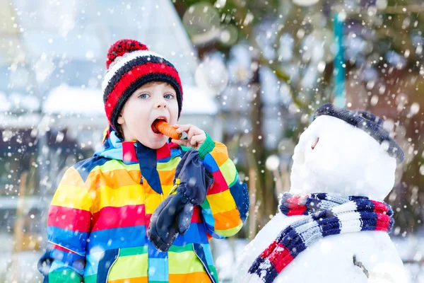 Ragazzo divertente in vestiti colorati facendo un pupazzo di neve, all'aperto — Foto Stock