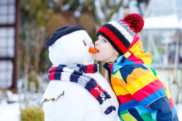 Αστείο παιδί αγόρι με πολύχρωμα ρούχα που κάνουν έναν χιονάνθρωπο, σε εξωτερικούς χώρους — Φωτογραφία Αρχείου