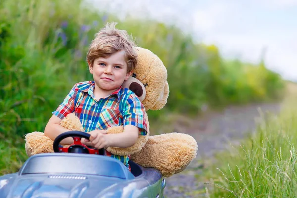 Niño pequeño conduciendo un coche de juguete grande con un oso, al aire libre . — Foto de Stock