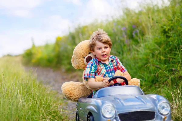 Kleiner Junge fährt großes Spielzeugauto mit einem Bären im Freien. — Stockfoto