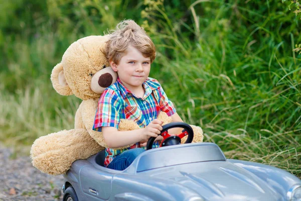 Niño pequeño conduciendo un coche de juguete grande con un oso, al aire libre . —  Fotos de Stock