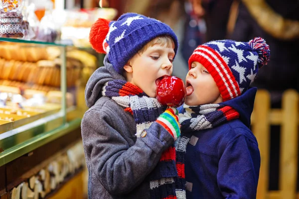 Dos niños pequeños comiendo azúcar manzana dulces de pie en el mercado de Navidad —  Fotos de Stock