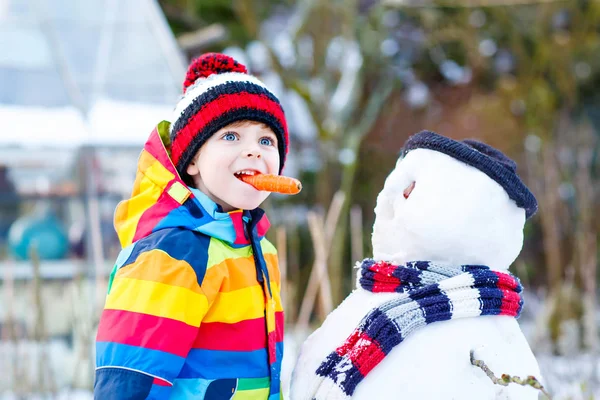 Funny kid boy in colorful clothes making a snowman, outdoors — Stock Photo, Image