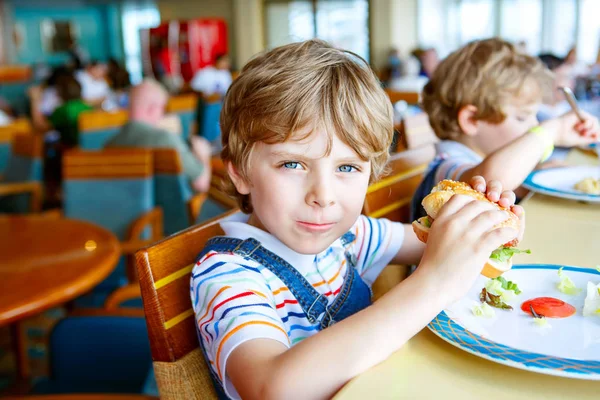 Lindo niño preescolar saludable come hamburguesa sentada en el comedor de la escuela —  Fotos de Stock