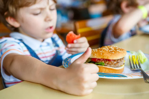 Ładny chłopak przedszkola zjada hamburger siedzi w stołówce szkolnej — Zdjęcie stockowe