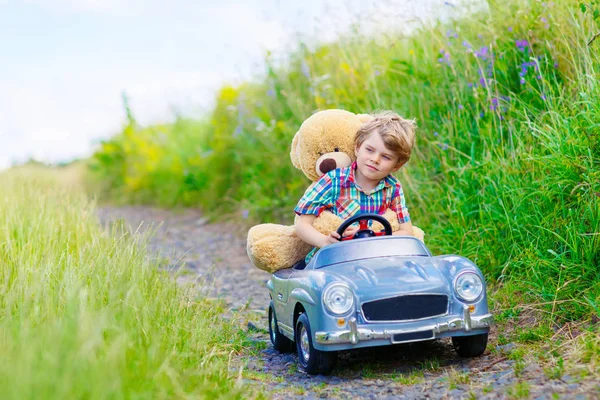 Kleiner Junge fährt großes Spielzeugauto mit einem Bären im Freien. — Stockfoto