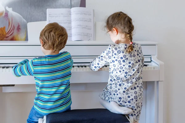 To små barnejenter og en gutt som spiller piano i stua eller på musikkskolen – stockfoto