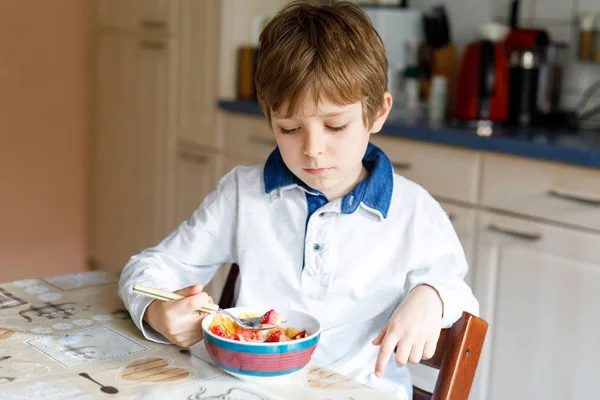 Pequeno menino da escola loira comendo cereais com leite e bagas, morango fresco para o café da manhã — Fotografia de Stock