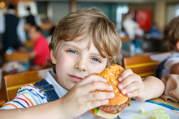 Ładny chłopak przedszkola zjada hamburger siedzi w stołówce szkolnej — Zdjęcie stockowe