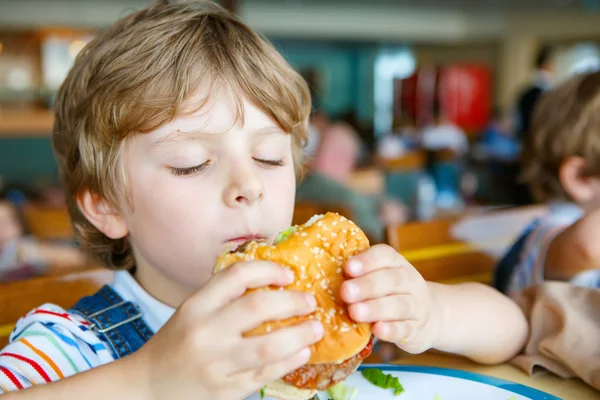 Lindo niño preescolar saludable come hamburguesa sentada en el comedor de la escuela —  Fotos de Stock